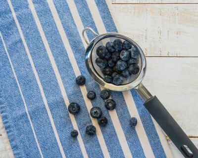 Fresh blueberries spilling from a small metal strainer onto a striped blue towel, highlighting antioxidant-rich, whole-food nutrition.