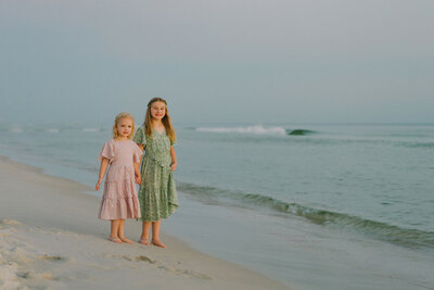 Two young girls stand barefoot on a sandy beach, wearing floral dresses. They hold hands, smiling with gentle ocean waves and an overcast sky behind them.