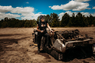Fierce woman in black warrior outfit poses beside a rusted car in a dusty field during a bold, Mad Max-inspired photoshoot.