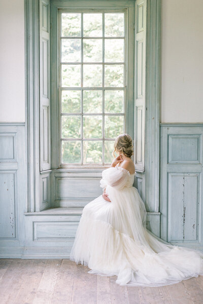 A pregnant mother wearing a long, flowy white gown sits in a window seat with a tall window, looking out while cradling her belly during her maternity session photographed by Newborn Photographer Baltimore Marie Elizabeth Photography