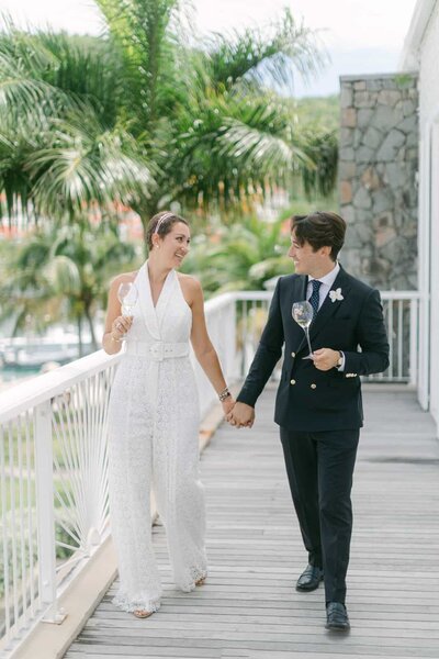 Elegant couple holding hands and smiling, walking through a palm-lined terrace in St Barth — Destination Wedding Photographer Portfolio.
