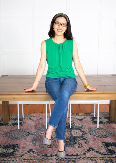 An Asian woman wearing a green sleeveless shirt and sitting on a table smiling.