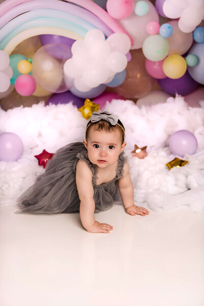 Baby girl in charcoal tulle gown crawls through whimsical rainbow and cloud themed first birthday portrait setup