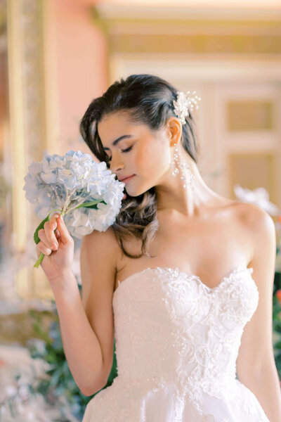 closeup of a bride smelling a blue hydrangea wearing a corseted wedding dress with the ballroom of avington park in the background