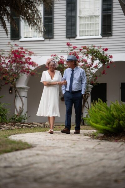 Elderly couple in wedding attire walking arm-in-arm on a stone pathway in front of a historic white colonial building with green shutters and blooming bougainvillea, likely after their St. Augustine courthouse wedding ceremony