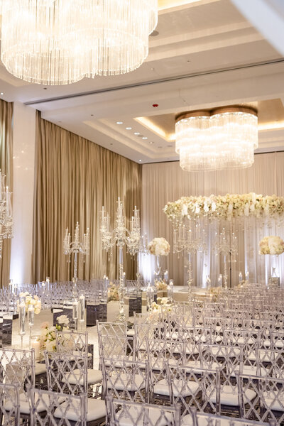 Grand ceremony with clear acrylic chairs, white and green florals and chandeliers up top. Greenish gold draping and white draping along walls and clear alter with white florals handing on top. 