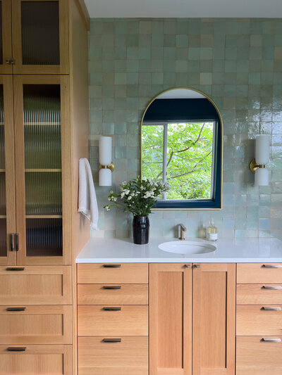 Large rustic wood vanity with lots of drawers, and dark grey countertops. Wall behind the vanity is off-white shiplap with a large arched mirror and two oil rubbed bronze sconces on either side. A widespread oil rubbed bronze faucet at the sink with a large beige vase with green florals to the left of the sink. To the right of the sink a jar of hand soap and a small beige dish. Large rustic black square floor tiles with brown grout.