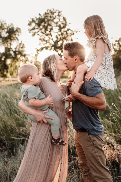 mom and dad kissing with daughter on dad's shoulders and baby boy on mom's hip