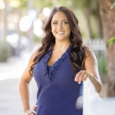 A realtor with REMI realty stands outdoors, smiling confidently for her professional branding photography session. One hand rests on a white fence, with trees and a blurred sidewalk in the background.