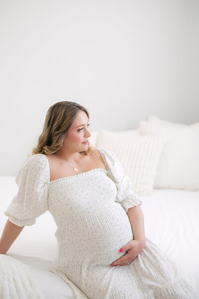 Photo of maternity mom sitting on a white bed holding her pregnant belly taken by newborn photographer missy marshall photography