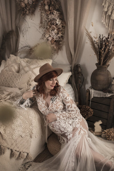 Femme enceinte assise près d’un lit en décor bohème, robe en dentelle blanche, chapeau en feutre, ambiance naturelle et douce.