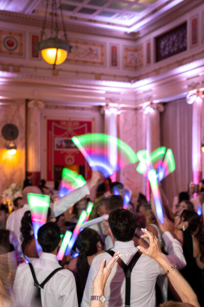 A lively wedding reception in a grand hall with elegant decor and intricate ceiling details. Guests are holding colorful light sticks, creating dynamic trails as they dance energetically. The atmosphere is festive and joyful.