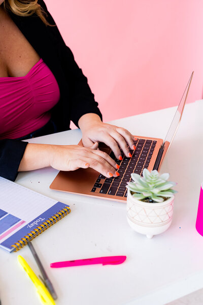 Woman typing on a rose gold apple laptop on a white desk with a light pink backdrop