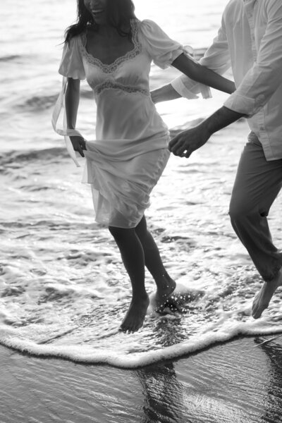 Couple running through the shallow water during their engagement session at Laguna Beach, captured in black and white, with waves splashing around them and a timeless, romantic feel.