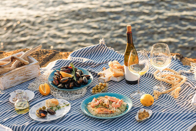 A seaside picnic spread on a blue and white striped cloth features plates of shrimp and rice, mussels, and olives. A bottle of wine and two glasses are set alongside, with a lemon and sliced bread completing the scene by the water.