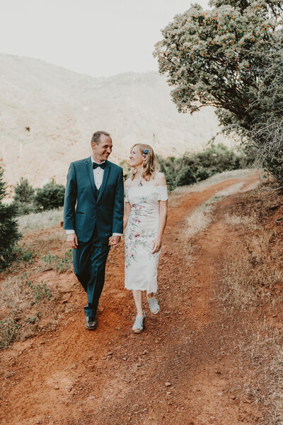 Bride wearing gown with flowers printed on it holding  hands with groom walking down trail looking at each other by Yosemite elopement photographer Kasey Mantiply
