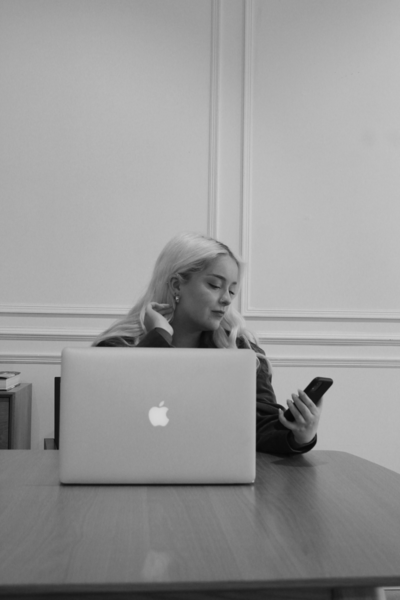 Black and white image of a woman holding an iphone and looking down at it. Her laptop is in the forefront and her other hand is pulling her hair behind her ear.