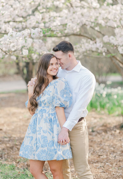 Engaged couple wearing white and blue holding hands and leaning on eachother in cherry blossoms