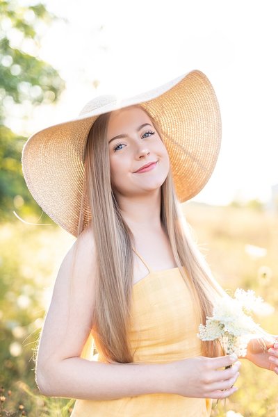 Senior wearing yellow dress and floppy light brown hat holding white wildflowers.