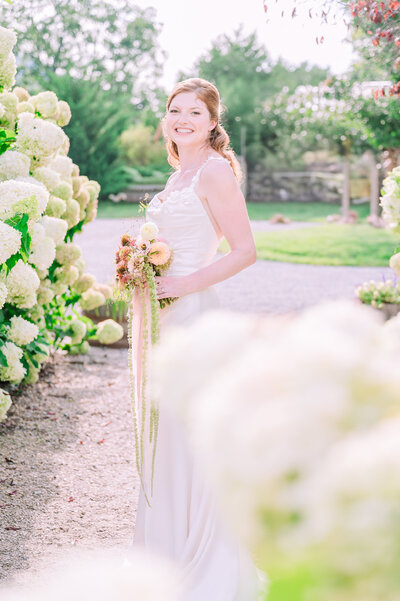 A smiling redheaded surrounded by white hydrangeas in greenery in the  North Carolina Blue Ridge Mountains on her wedding day by JoLynn Photography