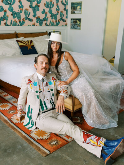 bride sits on hotel bed above groom on the floor in a printed bespoke suit looking at the camera without smiles