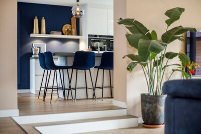 Luxe open keuken met donkerblauwe wand, witte kasten en fluwelen barstoelen met goudaccenten