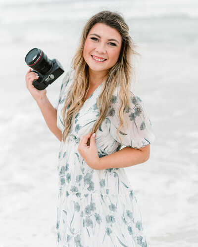 Abby, the owner of Abby Bell Photography, fine art wedding and portrait photographer in Alabama, smiling on the beach while holding her camera and wearing a floral dress.