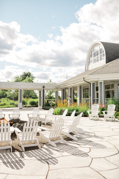 White Adirondack chairs circle a fire pit on a stone patio outside the Cliff House Maine, surrounded by flowers and coastal landscaping.