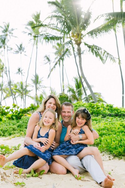 A family with two young girls sit on a beach with green palms behind them.