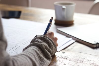 A close shot of a woman's arm in a light brown sweater. She has a blue ballpoint pen in her hand and is writing on a light wood table. Just in front of her is a white mug for coffee or tea.
