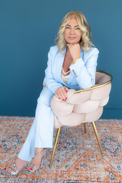 Personal development coach specializing in mid-life crisis recovery, seated in a unique pink chair and dressed in a pastel blue outfit.