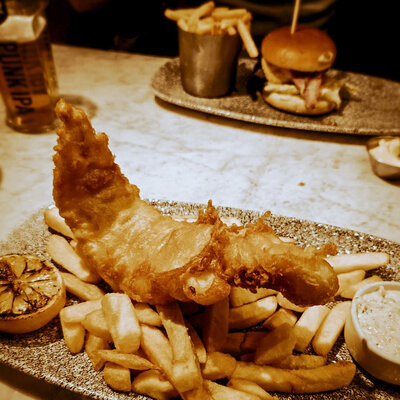 A plate of golden, crispy fish and chips is served with a charred lemon and a side of tartar sauce. In the background, another plate with a burger and fries can be seen, along with a bottle of beer on the table. The lighting gives the scene a warm, cozy feel.