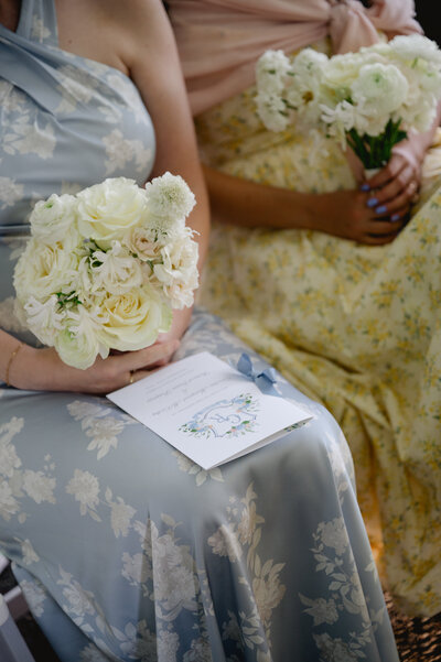 A woman seated in a floral patterned dress holding a white rose bouquet over her lap, with a wedding program placed on top of her skirt.
