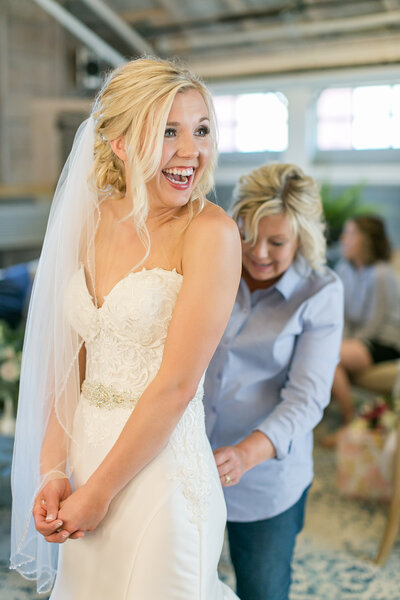 A white bride looking off to the side smiling brightly as another woman helps with her dress
