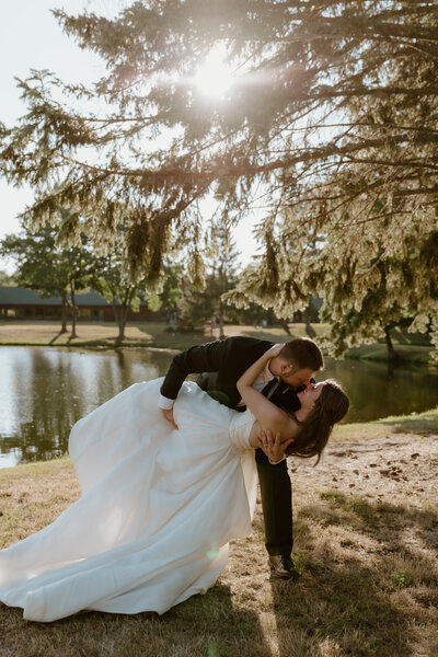 A couple shares a romantic kiss during a wedding photo shoot by a lake. The groom in a black suit dips the bride in a white gown under sunlight filtering through trees.