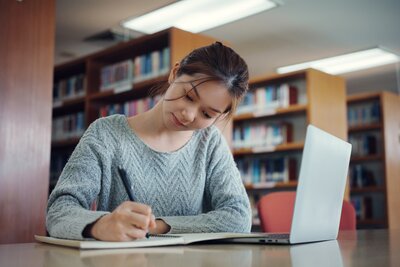 Asian woman in a blue sweater at s desk in a library looking down as she studies for her PA exams