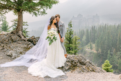 Bride and groom embracing at Surprise Corner in Banff National Park with the bride looking over her shoulder holding a bouquet and the Fairmont Banff Springs Hotel visible in the background