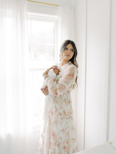 a mother stands at the window of her home holding her newborn on her chest