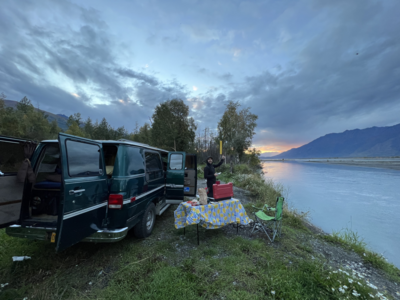 A green van is parked with it's doors open next to a river in Alaska, while Brian, an elopement photographer makes dinner next to the van.