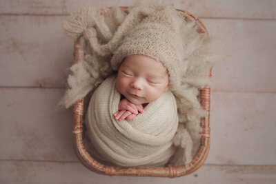a newborn wrapped in beige laying in a beautiful basket smiling
