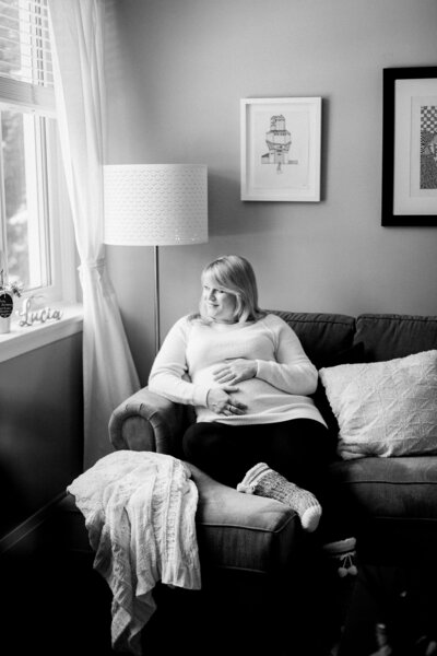 A black-and-white maternity portrait of a mother-to-be sitting in a cozy armchair, gazing thoughtfully. The intimate setting, with soft natural light coming through the window, captures a quiet and emotional moment of reflection during pregnancy. Photographed in a timeless, documentary style by KCP Families