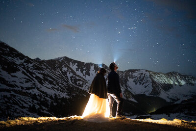 the couple is wearing a wedding dress and a tan suit. they are facing backwards towards a mountain peak. there is greenery surrounding them, and they are embracing each other while enjoying the view. they are eloping in with their dog and finished their hiking elopement in a valley surrounded by the san juan mountain range. they are reading their vows and signing their marriage license in the  mountains.