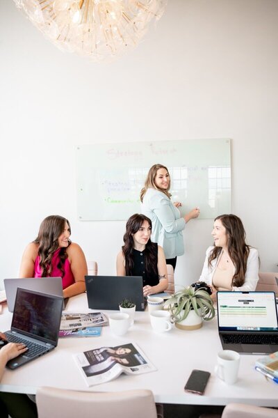 Pierce PR team members collaborating around a table with laptops, coffee, and magazines while one team member writes on a strategy board in a modern, well-lit office.