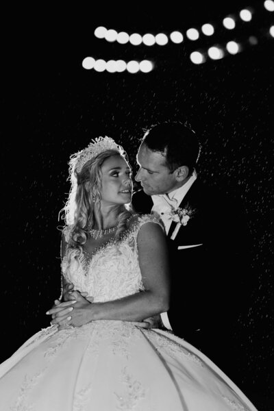 Emotive and luxurious black-and-white wedding portrait of a bride and groom holding each other under soft rain and romantic lights.