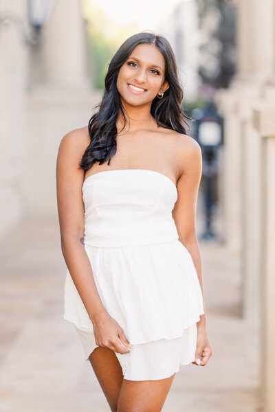 Elegant senior portrait of a young woman in a white dress, smiling confidently during a golden hour photo session at Adriatica Village in McKinney. Captured in a light and airy style by a DFW-based senior photographer.