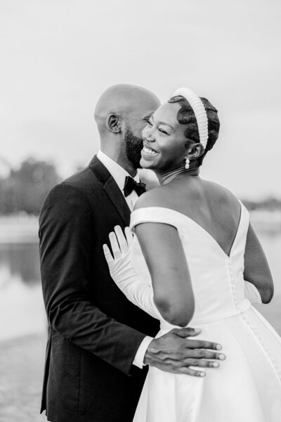 A joyful bride in an elegant off-the-shoulder gown shares a loving moment with her groom at their luxury wedding in Savannah, GA. A timeless black-and-white portrait.