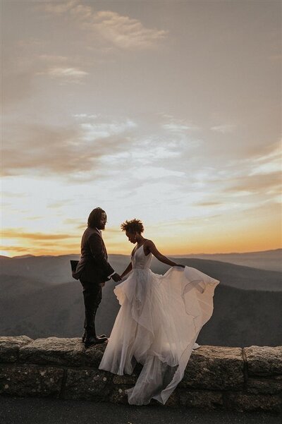 Two people hold hands on a scenic mountain overlook in Virginia's Blue Ridge Mountains, with one person’s wedding dress flowing in the wind at sunset.