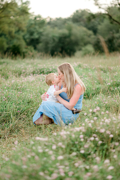 Photo of a mom kissing her toddler,  while standing in a field of flowers