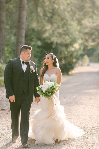 a man in a black tux holding hands with a bride in a white dress with a white bouquet walking  towards the camera and looking at one another