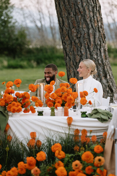 Bride and groom sitting at their head table, sharing a laugh, surrounded by tall floral arrangements.
